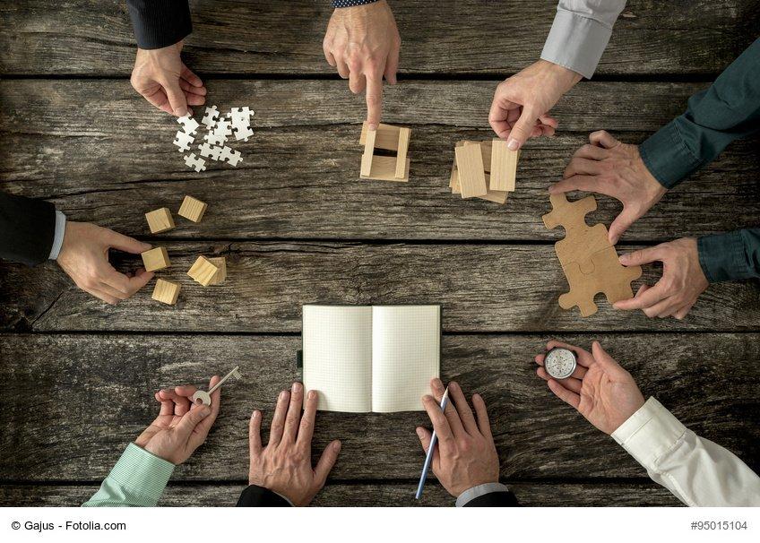 Eight businessmen planning a strategy in business advancement each holding different but equally important metaphorical element - compass, puzzle pieces, pegs, cubes, key and one making notes.