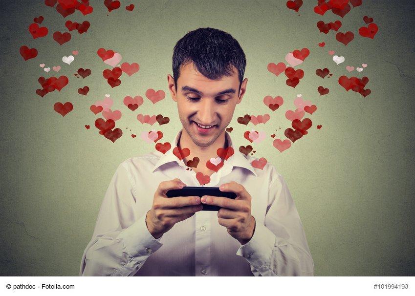 Portrait young handsome happy man sending receiving love sms text message on mobile phone with red hearts flying away up isolated on grey wall background. Human emotions