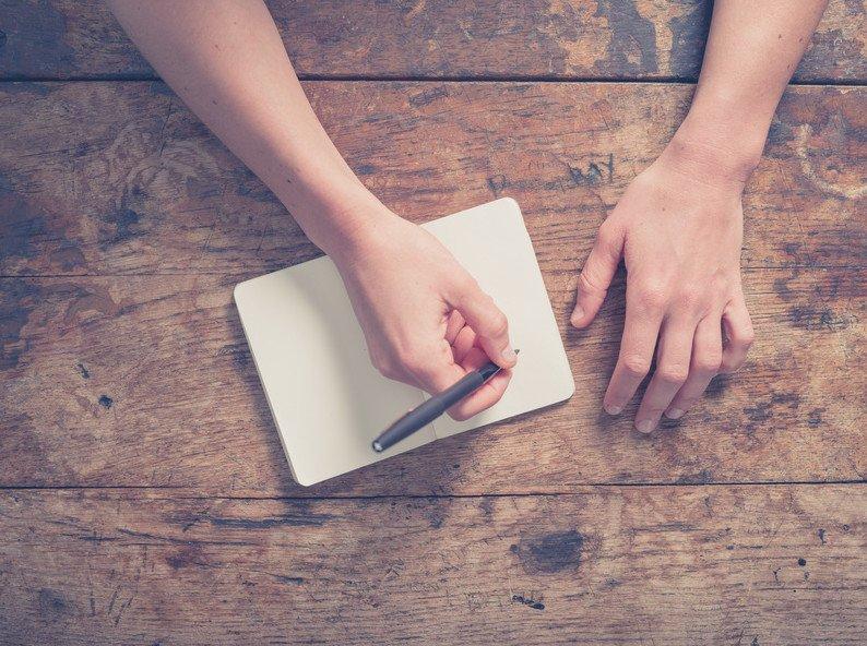 Close up on the hands of a young woman as she is writing in a small notepad at a wooden table