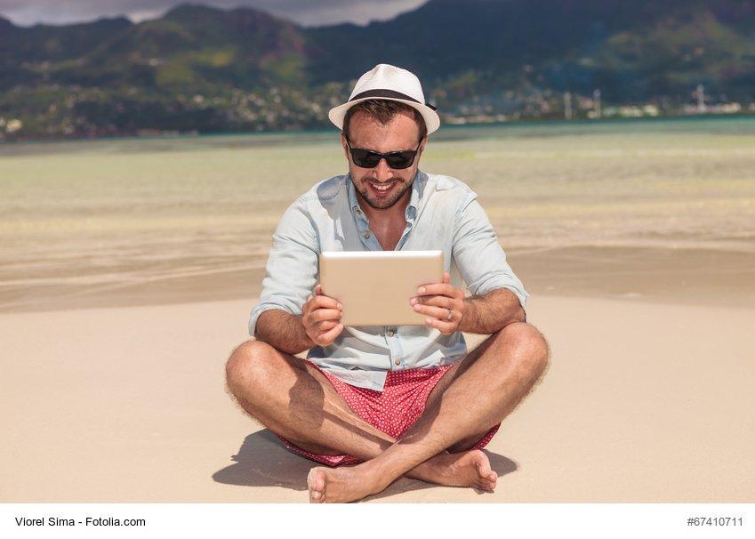 smiling man with sunlasses and hat reading on a tablet computer while sitting on the beach
