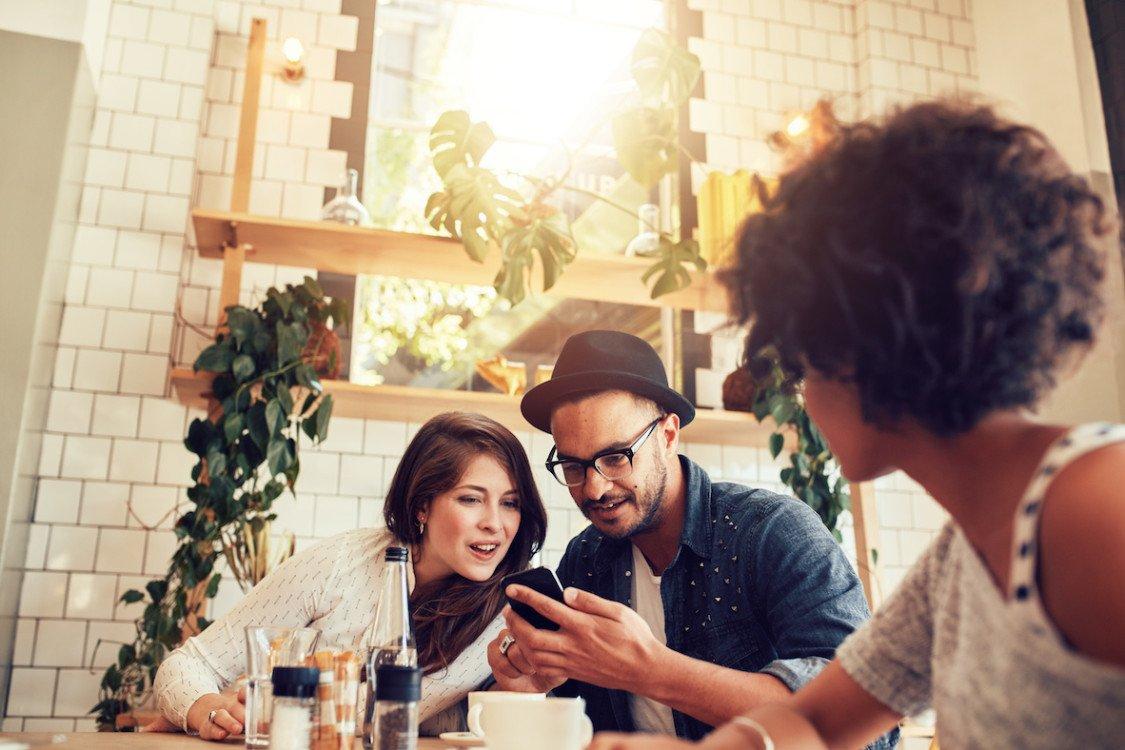 Young couple looking at smart phone while sitting in cafe with friends. Group of young people sitting at a table in restaurant and using mobile phone.