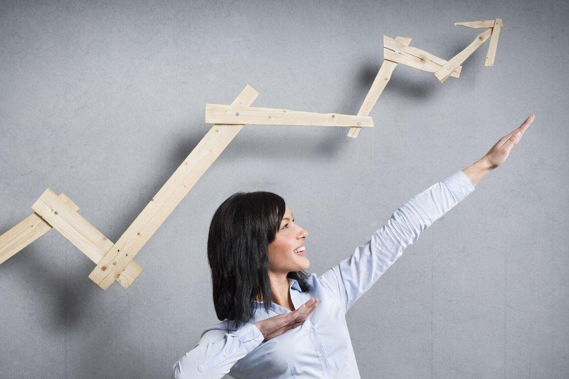 Concept: Successful business trend. Happy talented businesswoman pointing arm upwards in front of ascending business graph, isolated on grey background.