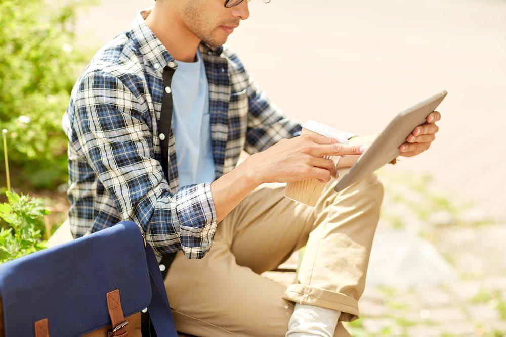 business, education, technology, communication and people concept - close up of creative or hipster man with tablet pc computer drinking coffee from paper cup and sitting on city street bench