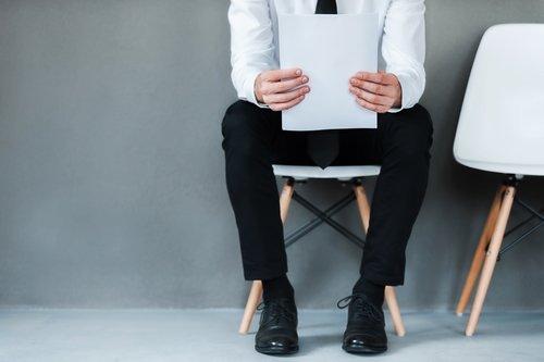 Waiting for interview. Close-up of young man holding paper while sitting on chair against grey background